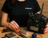 Siyah "makerion" tişört giymiş, kırmızı ojeli elleriyle RC verici, fırçasız motor, ESC, flight controller, LiPo batarya ve servo motor gibi donanımları ahşap yüzey üzerinde sergileyen kadın.