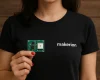 Makerion logolu siyah tişört giyen bir kadın, kırmızı ojeli parmaklarıyla Ultra Wideband (UWB) geliştirme kartı tutuyor; arka planda ahşap yüzey yer alıyor.